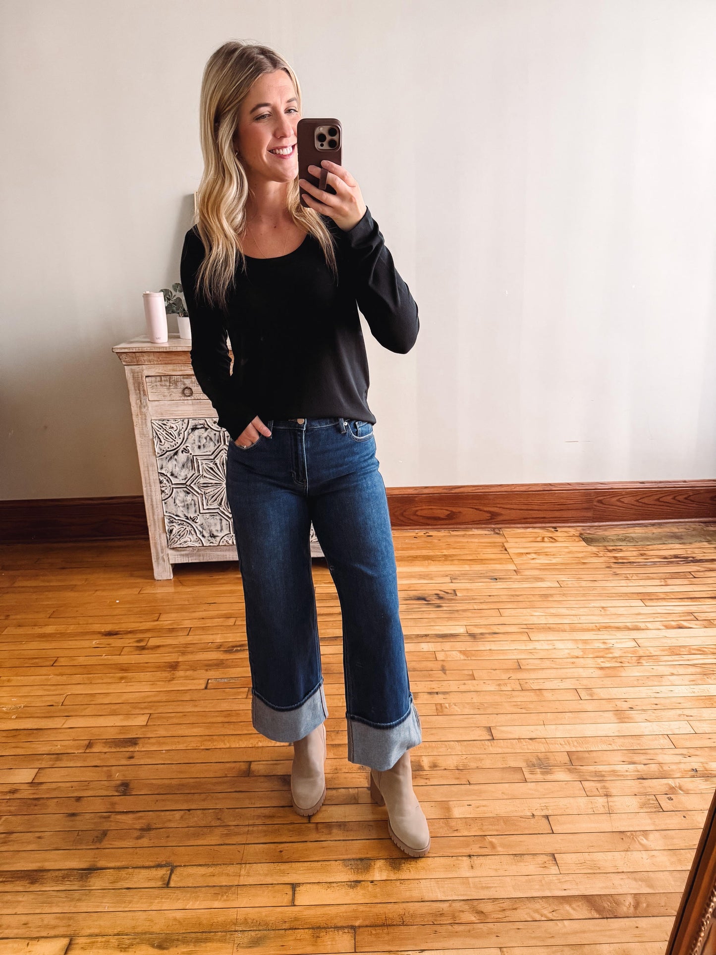 Woman taking a mirror selfie wearing a black top and blue jeans in a room with wooden flooring.