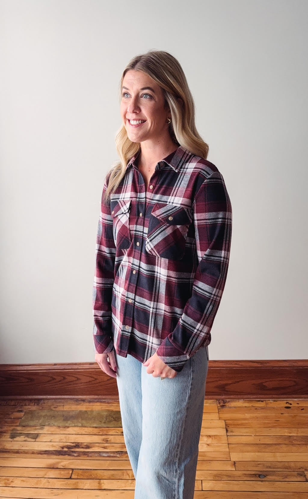 Woman wearing a plaid shirt and jeans standing on a wooden floor with a white wall background