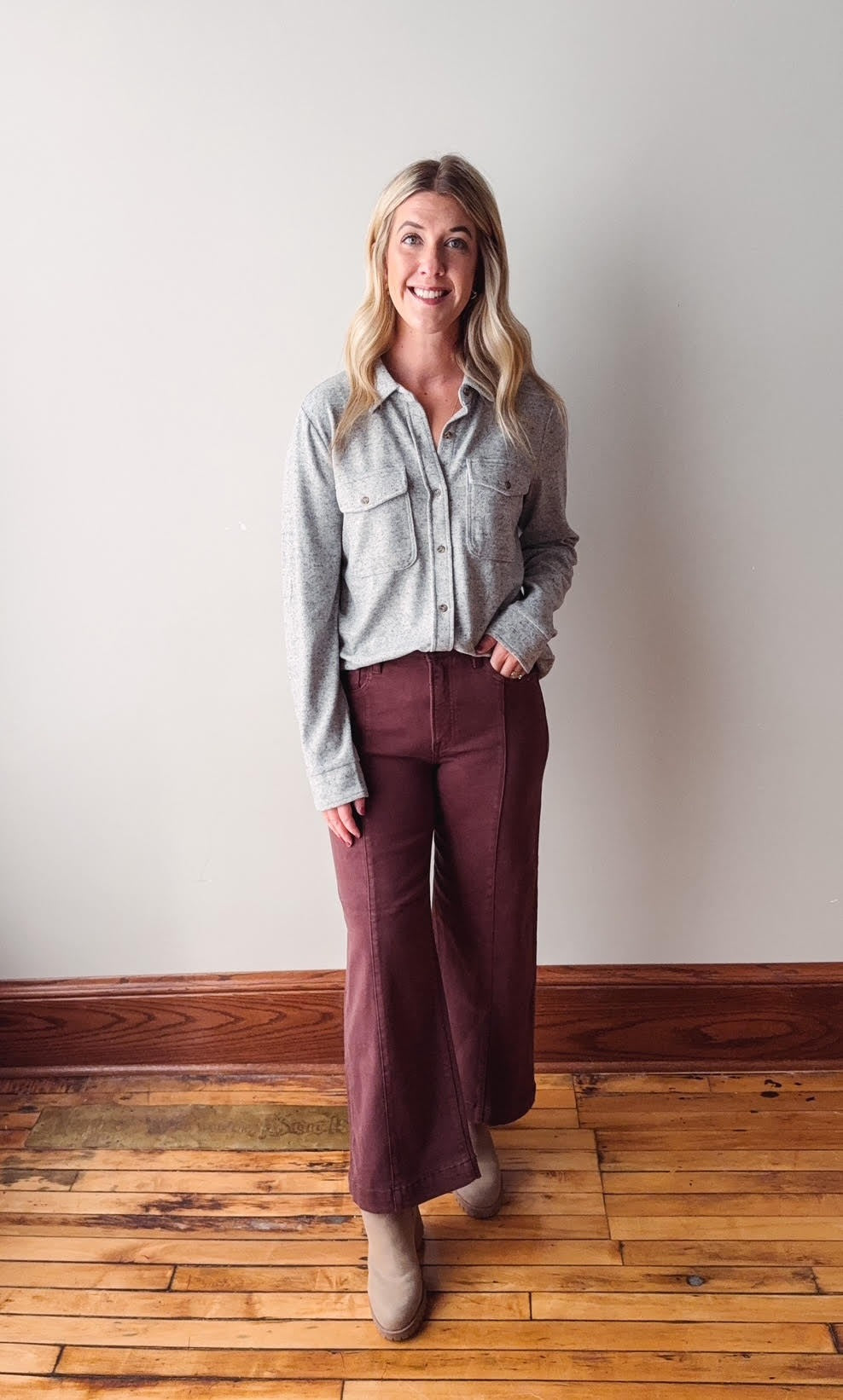 Woman wearing a light gray flannel shirt and burgundy pants standing on a wooden floor against a white wall.