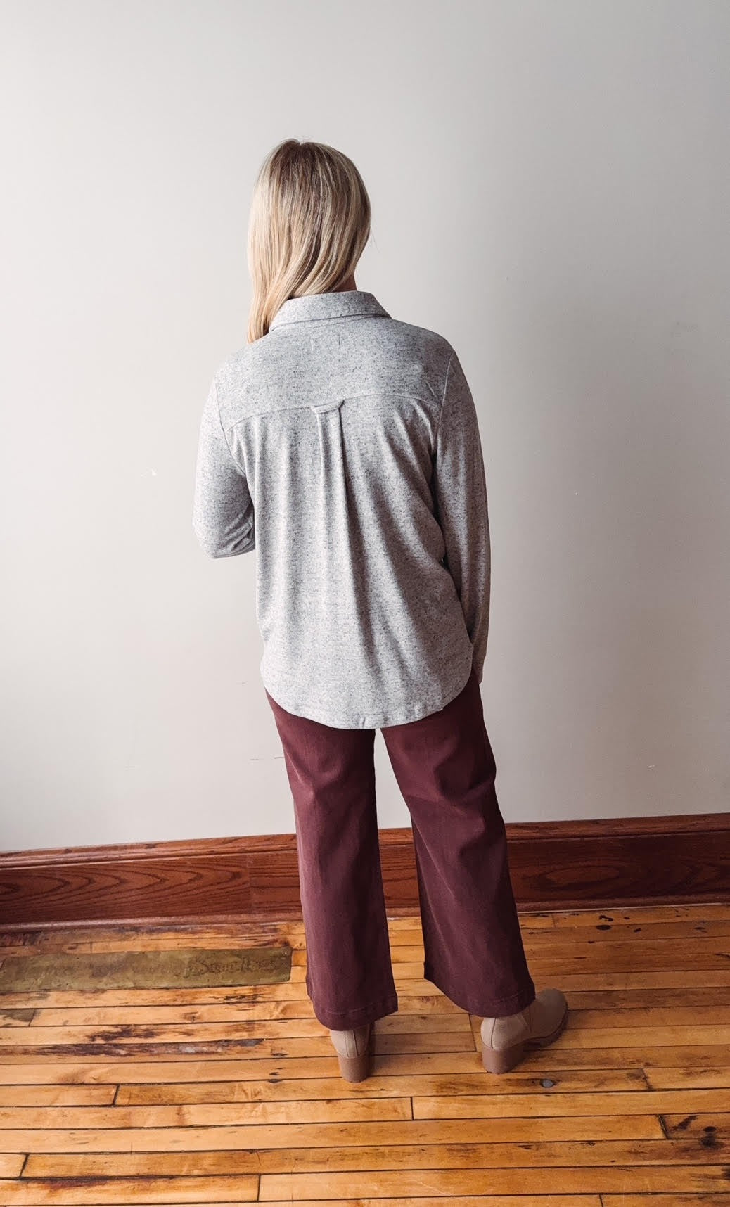 Person wearing a gray sweater and maroon pants standing in a room with wooden flooring and white walls.