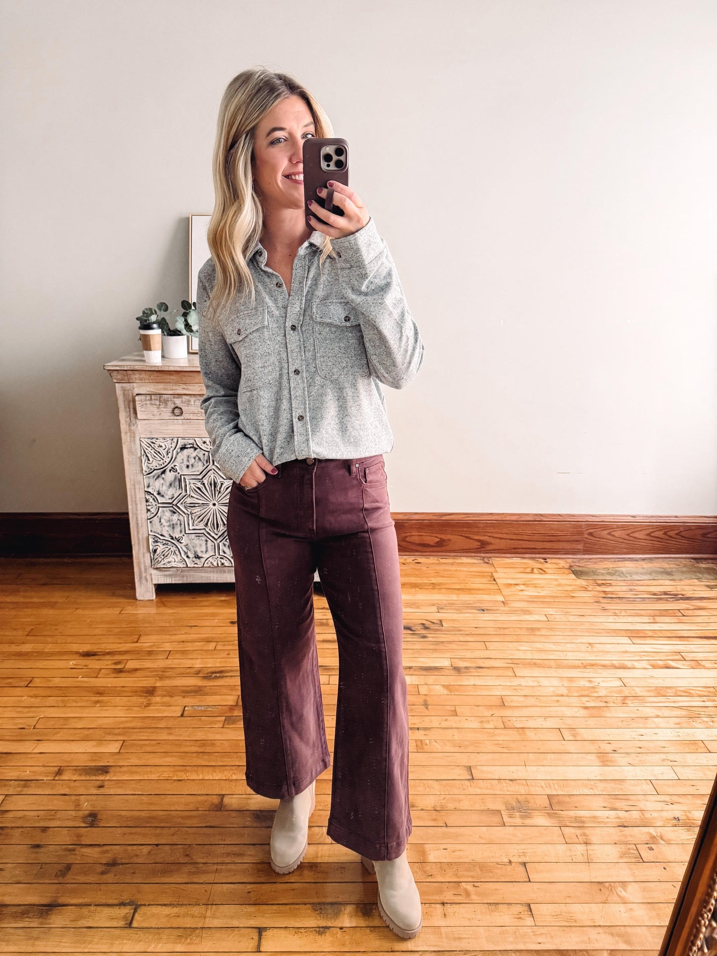 Woman taking a mirror selfie wearing a gray button-up shirt and purple pants in a room with wooden flooring.