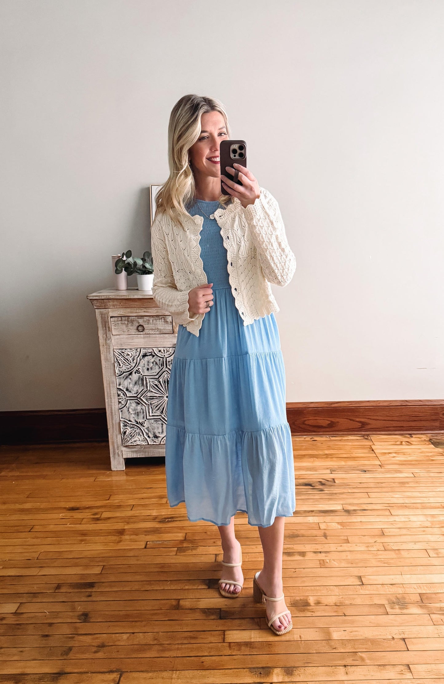 Woman taking a mirror selfie wearing a light blue dress and cream cardigan in a room with wooden flooring.
