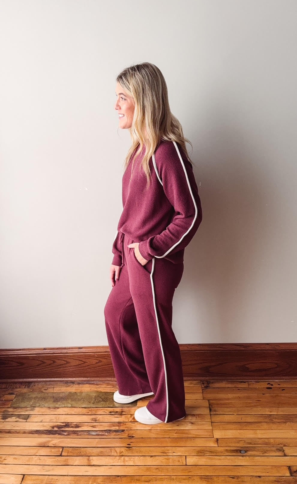 Woman wearing a burgundy tracksuit with white piping against a plain wall.