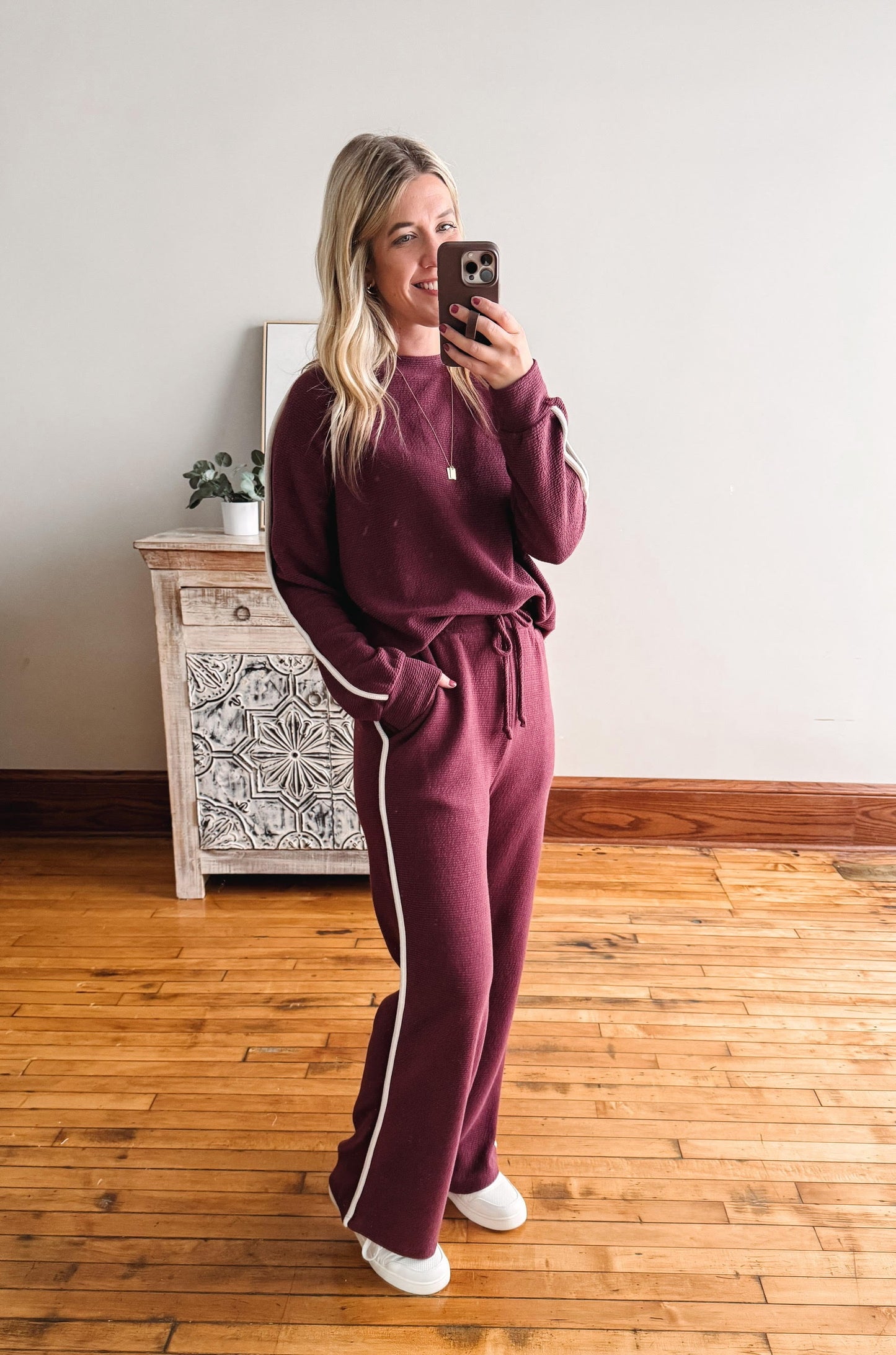 Woman taking a mirror selfie wearing a burgundy tracksuit with white stripes in a room with wooden flooring.