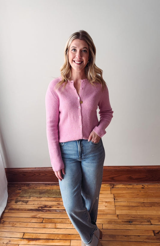 Woman wearing a pink sweater and blue jeans standing in a room with wooden flooring and white walls.