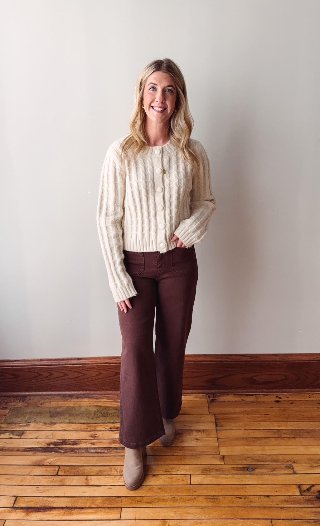 Woman wearing a cream sweater and brown pants standing on a wooden floor against a white wall.