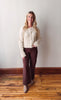 Woman wearing a cream sweater and brown pants standing on a wooden floor against a white wall.