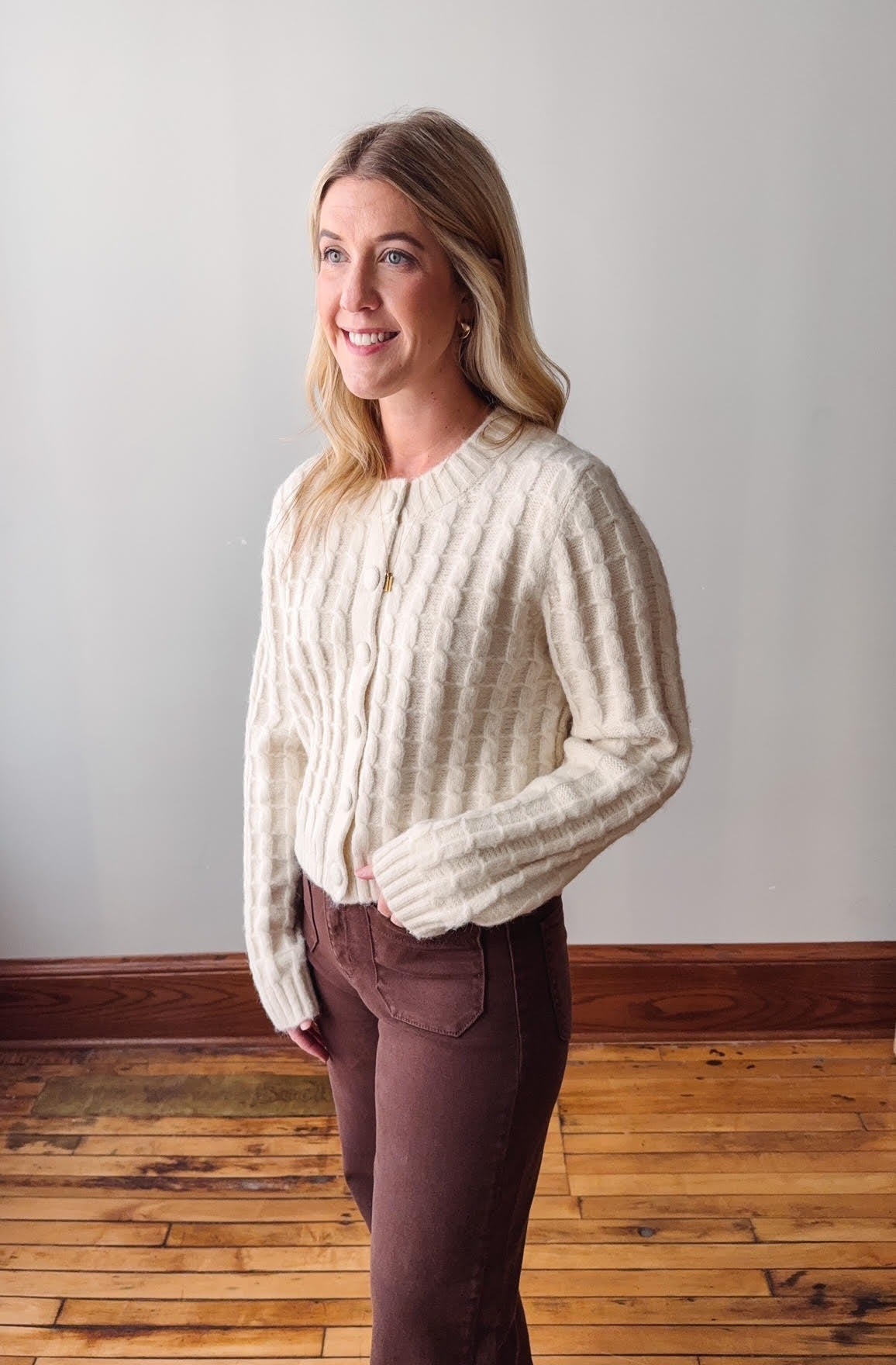 Woman wearing a cream cardigan and brown pants standing against a plain wall.