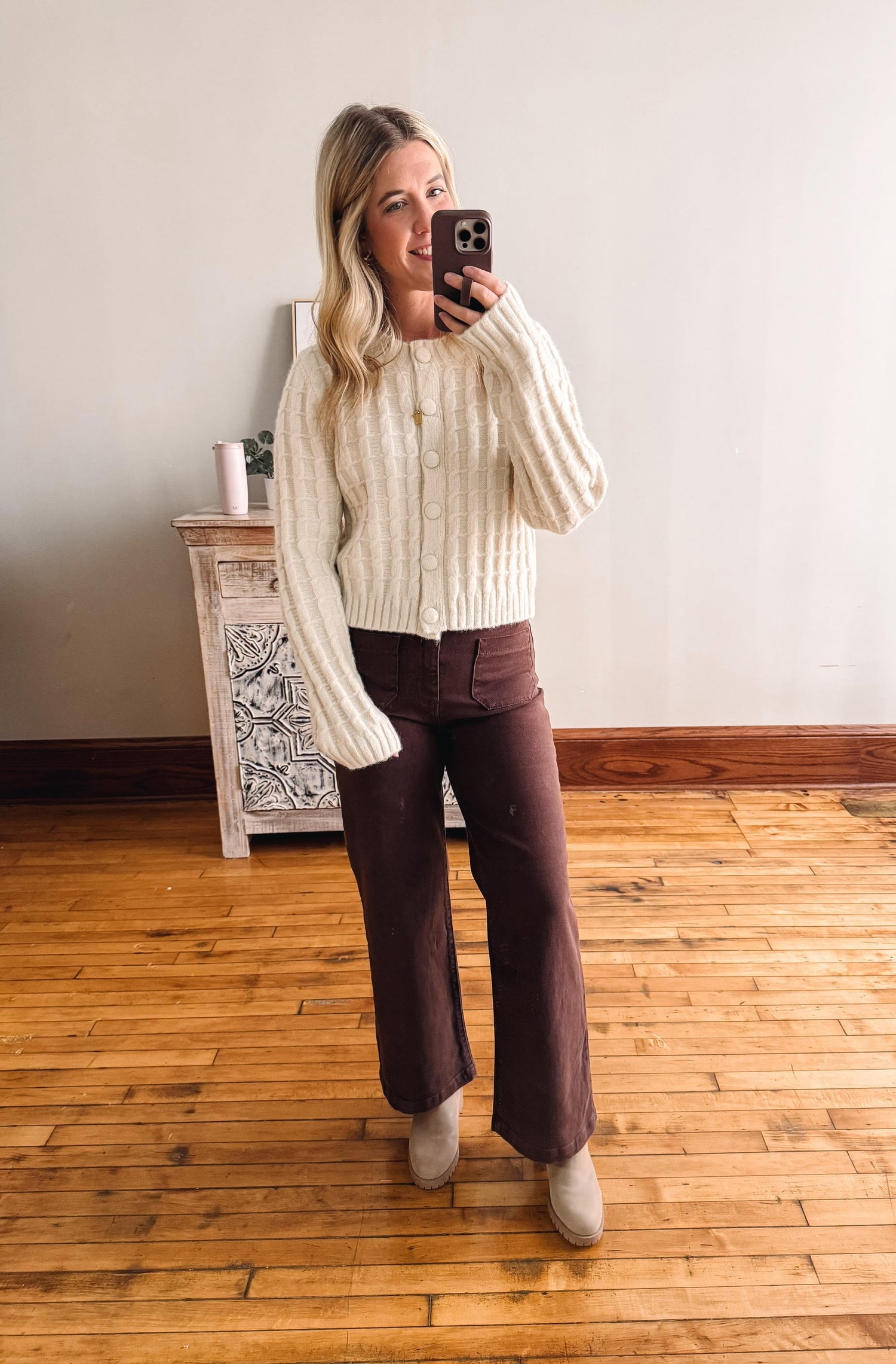 Woman taking a mirror selfie wearing a cream sweater and brown pants in a room with wooden flooring.
