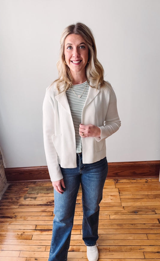 Woman wearing a white blazer, striped shirt, and jeans standing on a wooden floor.
