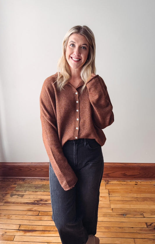 Woman wearing a brown sweater and dark jeans standing on a wooden floor with a white wall background