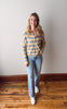 Woman wearing a striped sweater and jeans standing on a wooden floor against a white wall.