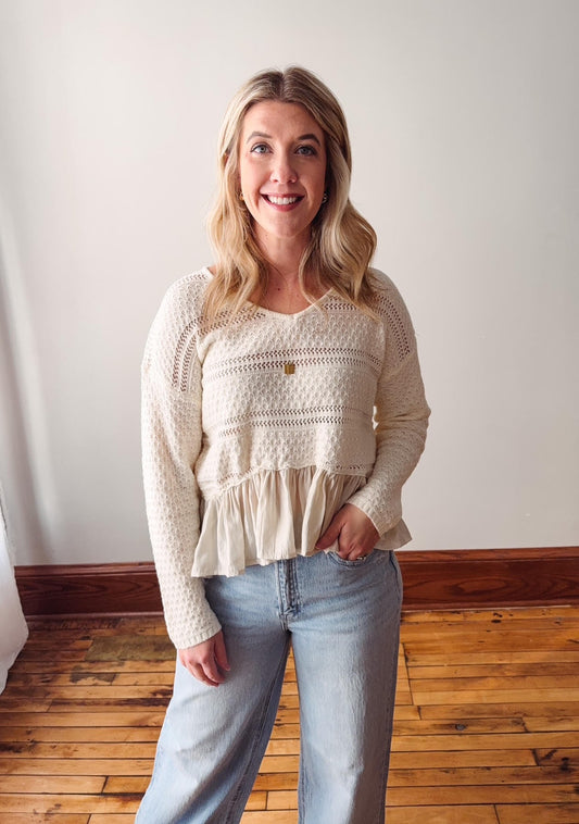 Woman wearing a white lace top and light blue jeans standing in a room with wooden flooring and a white wall.