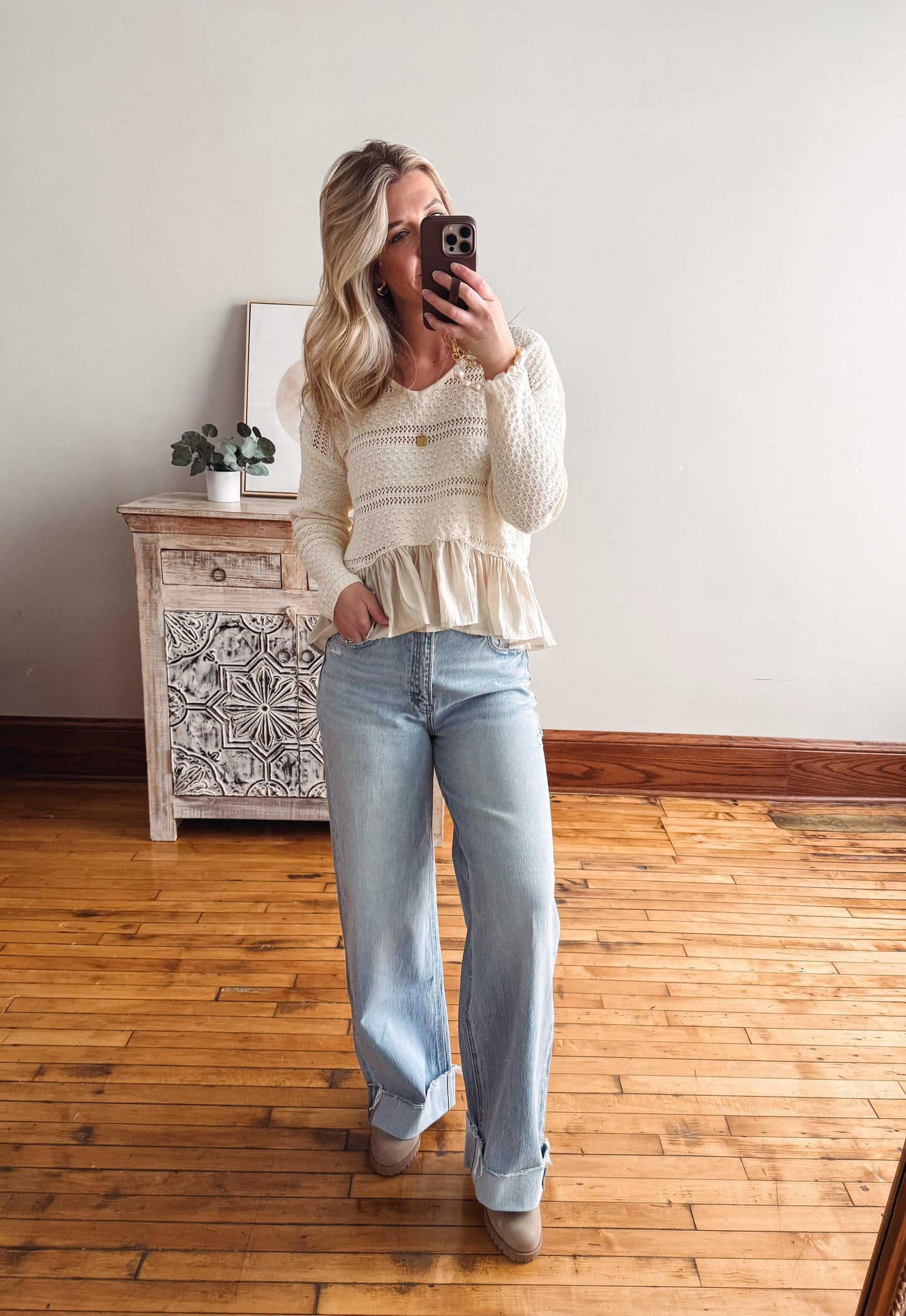 Person wearing a white lace top and light blue jeans in a room with wooden flooring and a decorative cabinet.