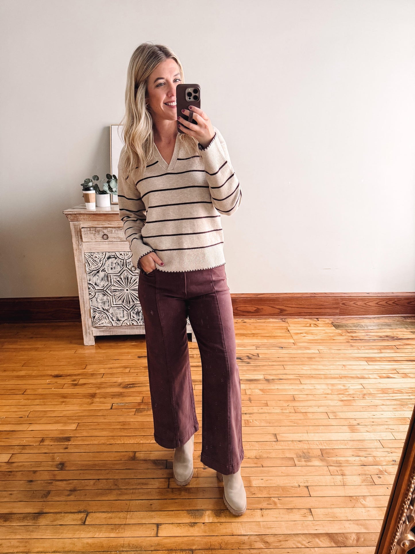 Woman taking a mirror selfie wearing a striped sweater and burgundy pants in a room with wooden flooring.