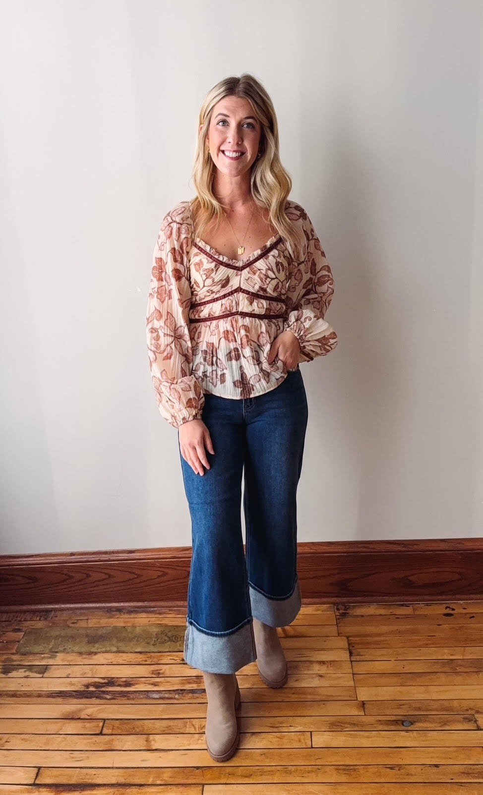 Woman wearing a patterned blouse and blue jeans standing on a wooden floor against a white wall.