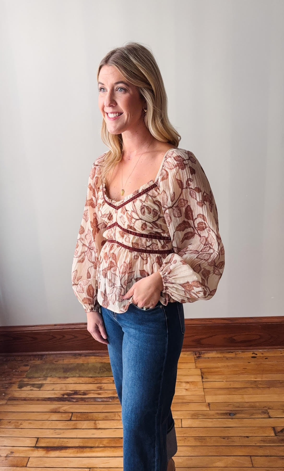 Woman wearing a floral blouse and blue jeans standing against a white wall.