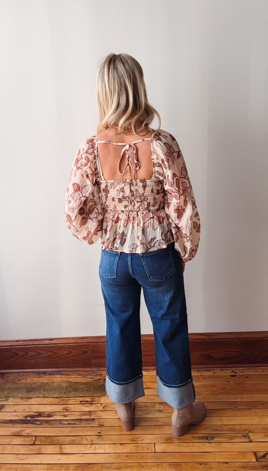 Person wearing a floral blouse and blue jeans standing against a plain wall.