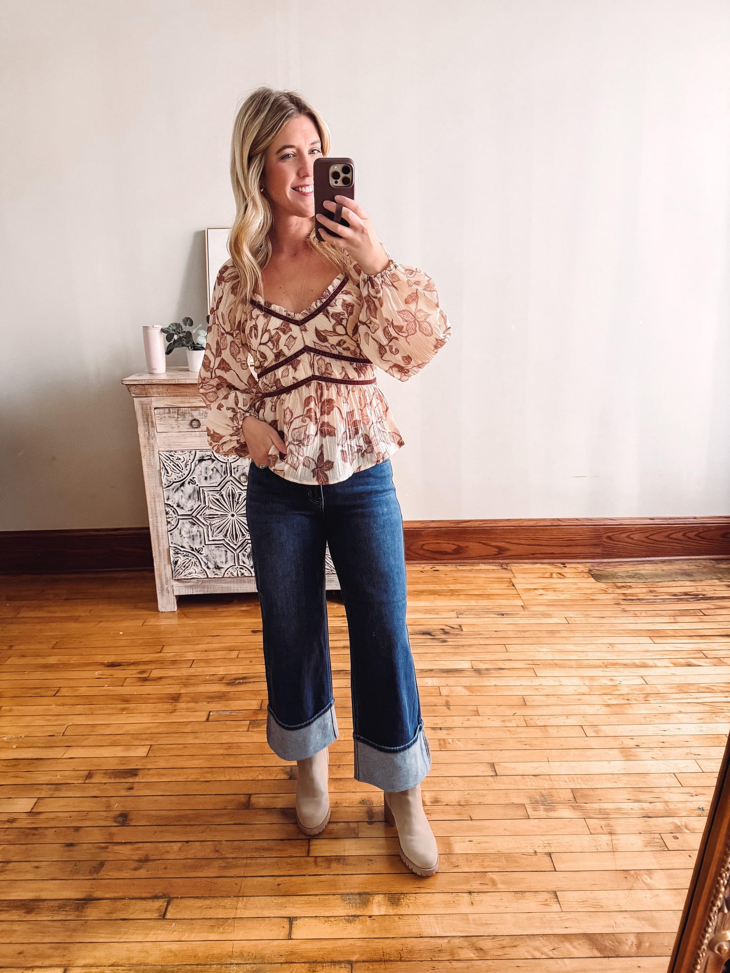 Woman taking a mirror selfie wearing a floral blouse and blue jeans in a room with wooden flooring.