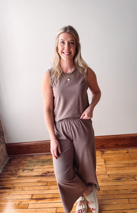 Woman wearing a brown sleeveless top and pants standing on a wooden floor.