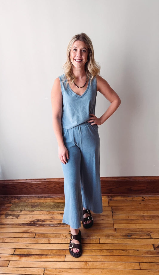 Woman wearing a light blue sleeveless top and wide-leg pants standing on a wooden floor.