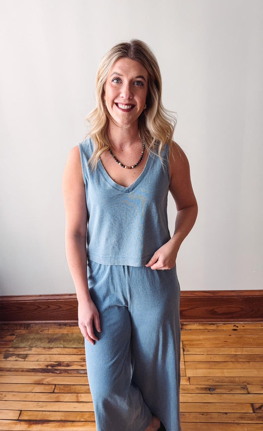 Woman wearing a light blue sleeveless top and pants standing against a plain wall.