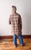 Person wearing a plaid coat and blue jeans standing on a wooden floor against a white wall.