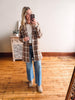 Woman taking a mirror selfie wearing a plaid coat, white shirt, blue jeans, and beige boots in a room with wooden flooring.