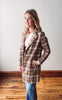 Woman wearing a plaid coat standing in a room with wooden flooring and a white wall.