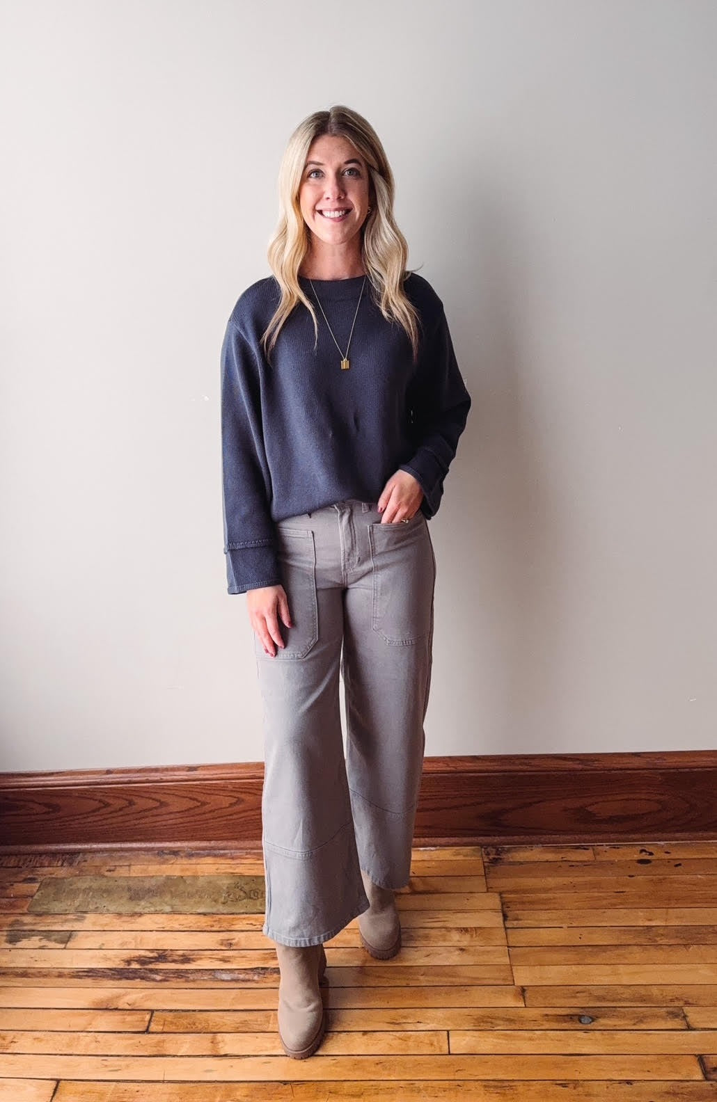 Woman wearing a navy sweater and gray pants standing against a plain wall.