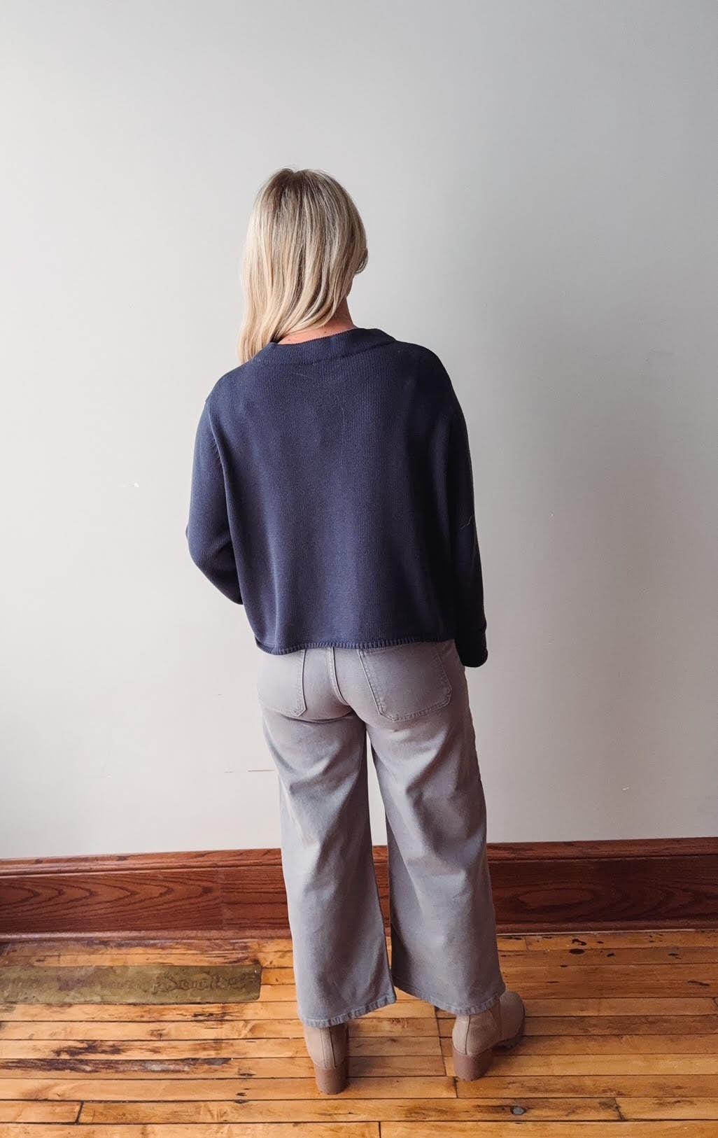 Person wearing a navy sweater and gray pants standing against a plain wall.