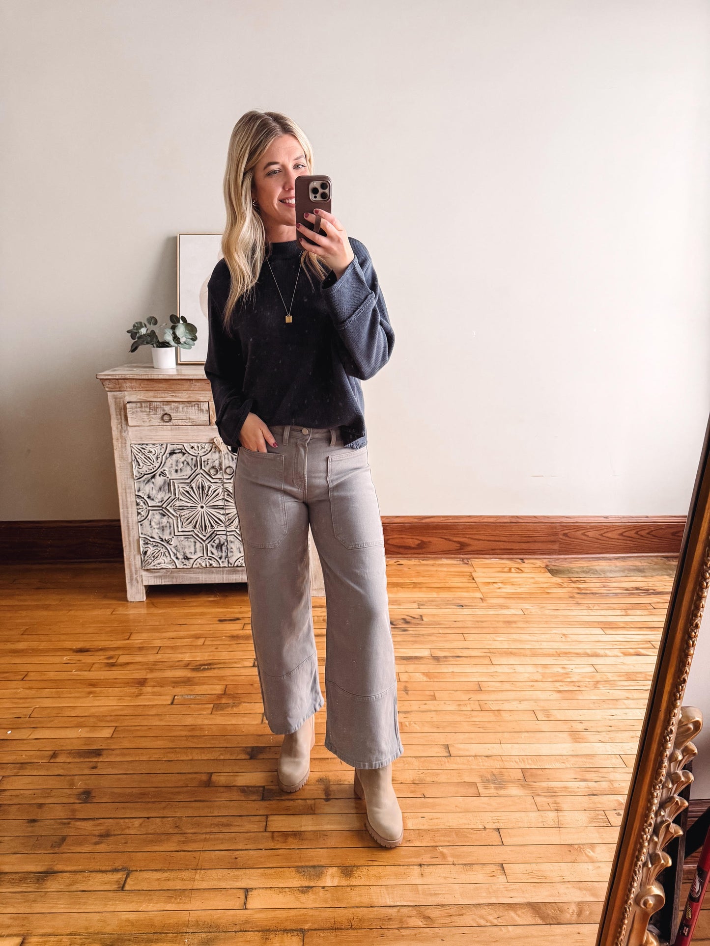 Woman taking a mirror selfie wearing a navy sweater and light gray jeans in a room with wooden flooring.