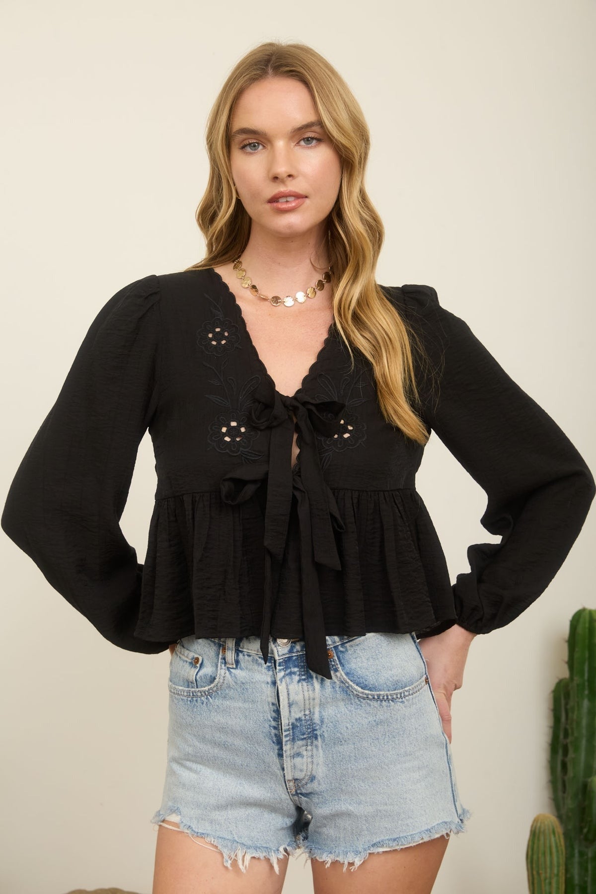 Woman wearing a black top with ruffled details and denim shorts against a neutral background.