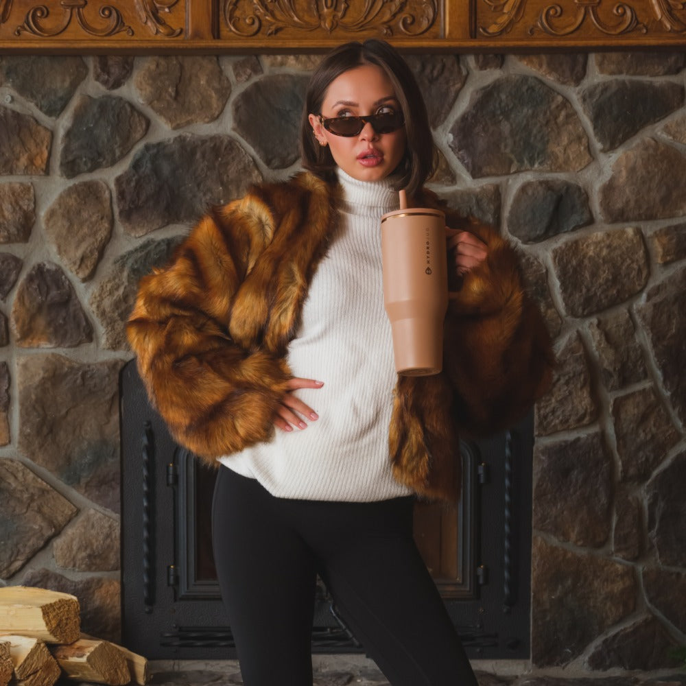 Person wearing a fur coat and sunglasses holding a pink cup against a stone fireplace background
