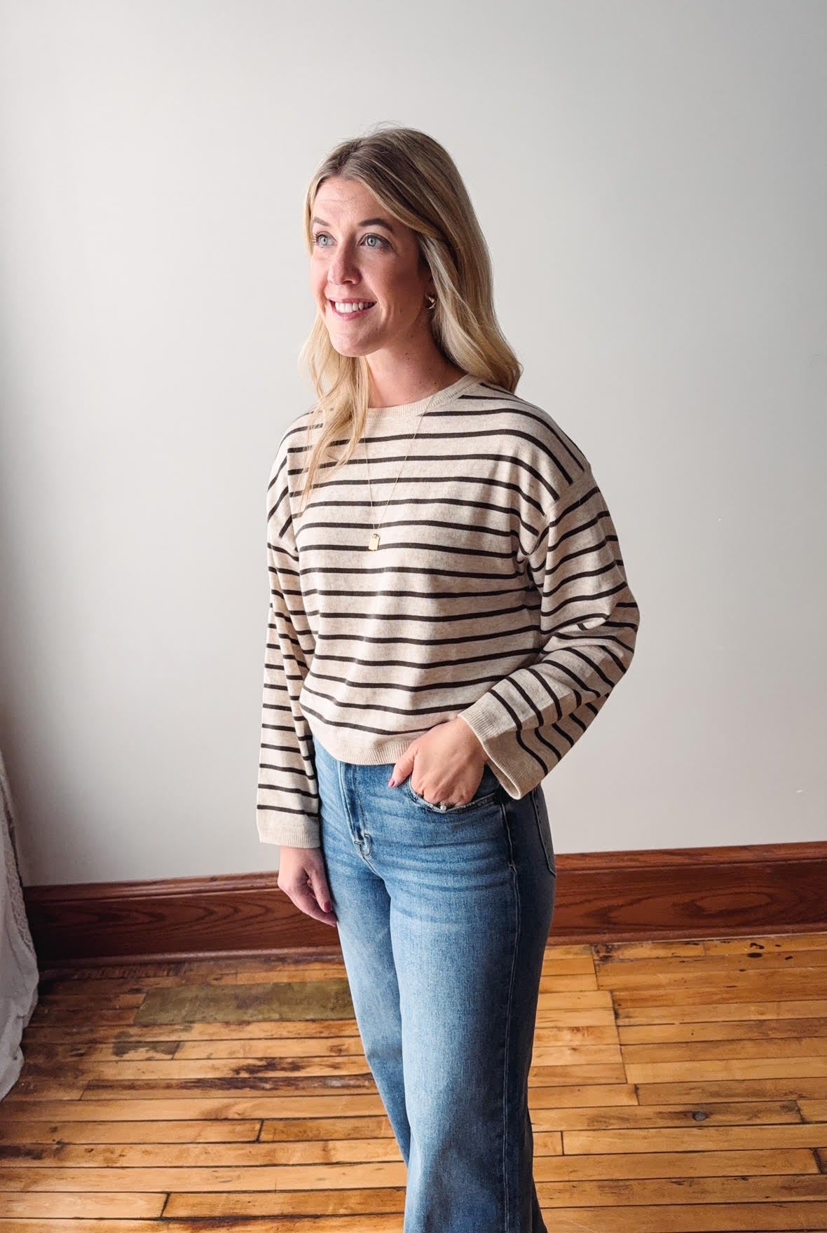 Woman wearing a striped sweater and jeans standing in a room with wooden flooring and a white wall.