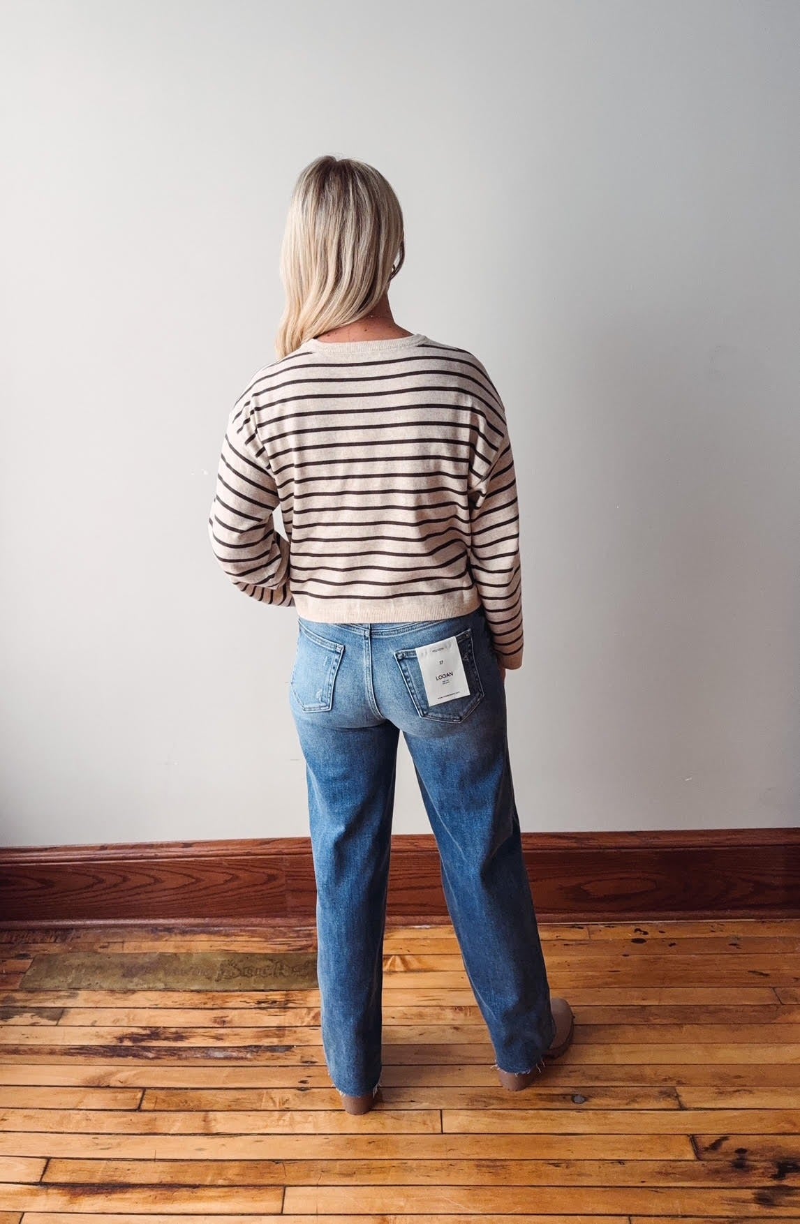Person wearing a striped sweater and blue jeans standing on a wooden floor with a white wall background