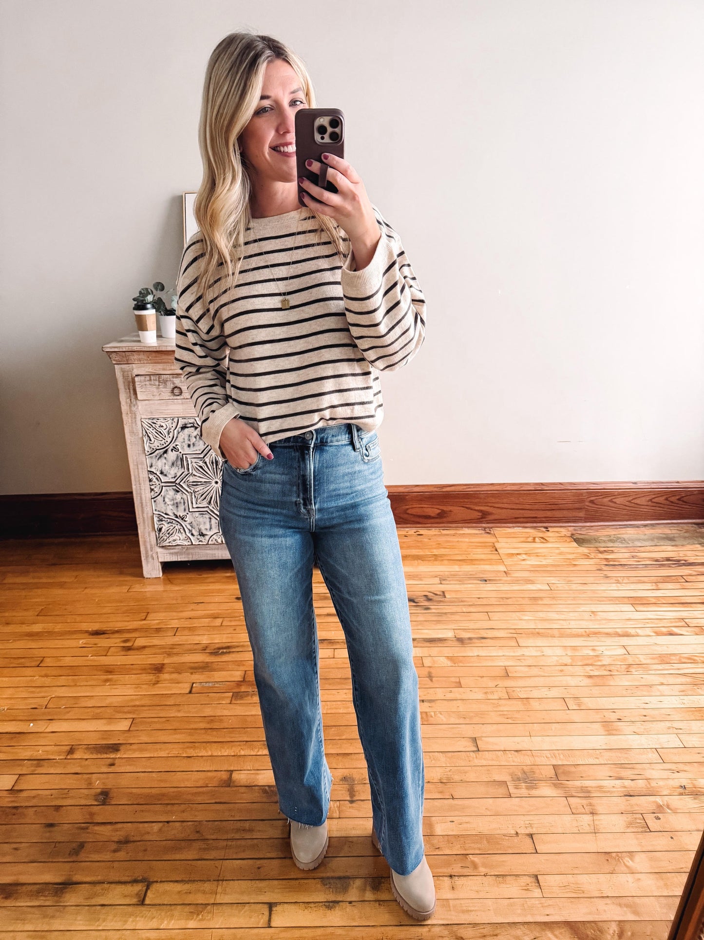 Woman taking a mirror selfie wearing a striped sweater and blue jeans in a room with wooden flooring.