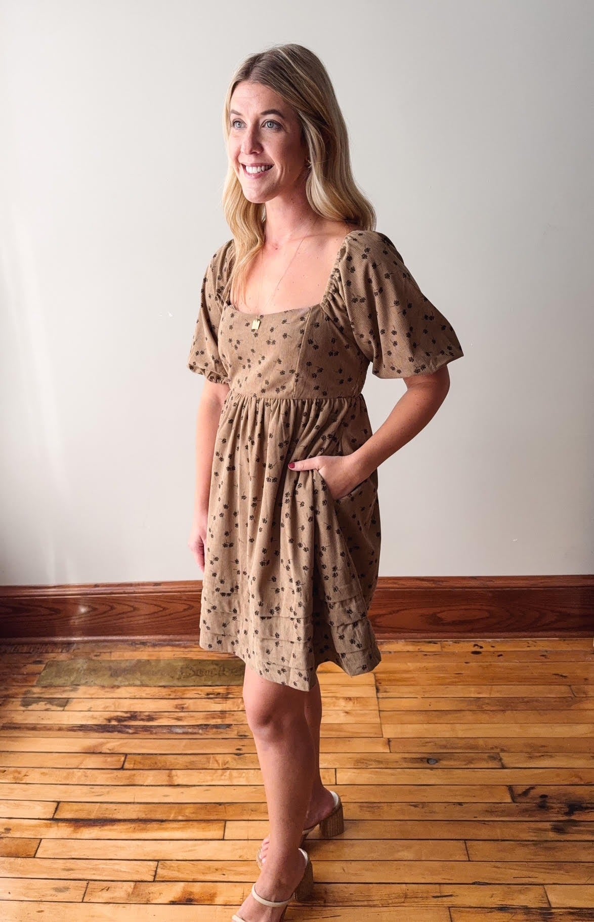 Woman wearing a patterned dress standing on a wooden floor with a white wall background