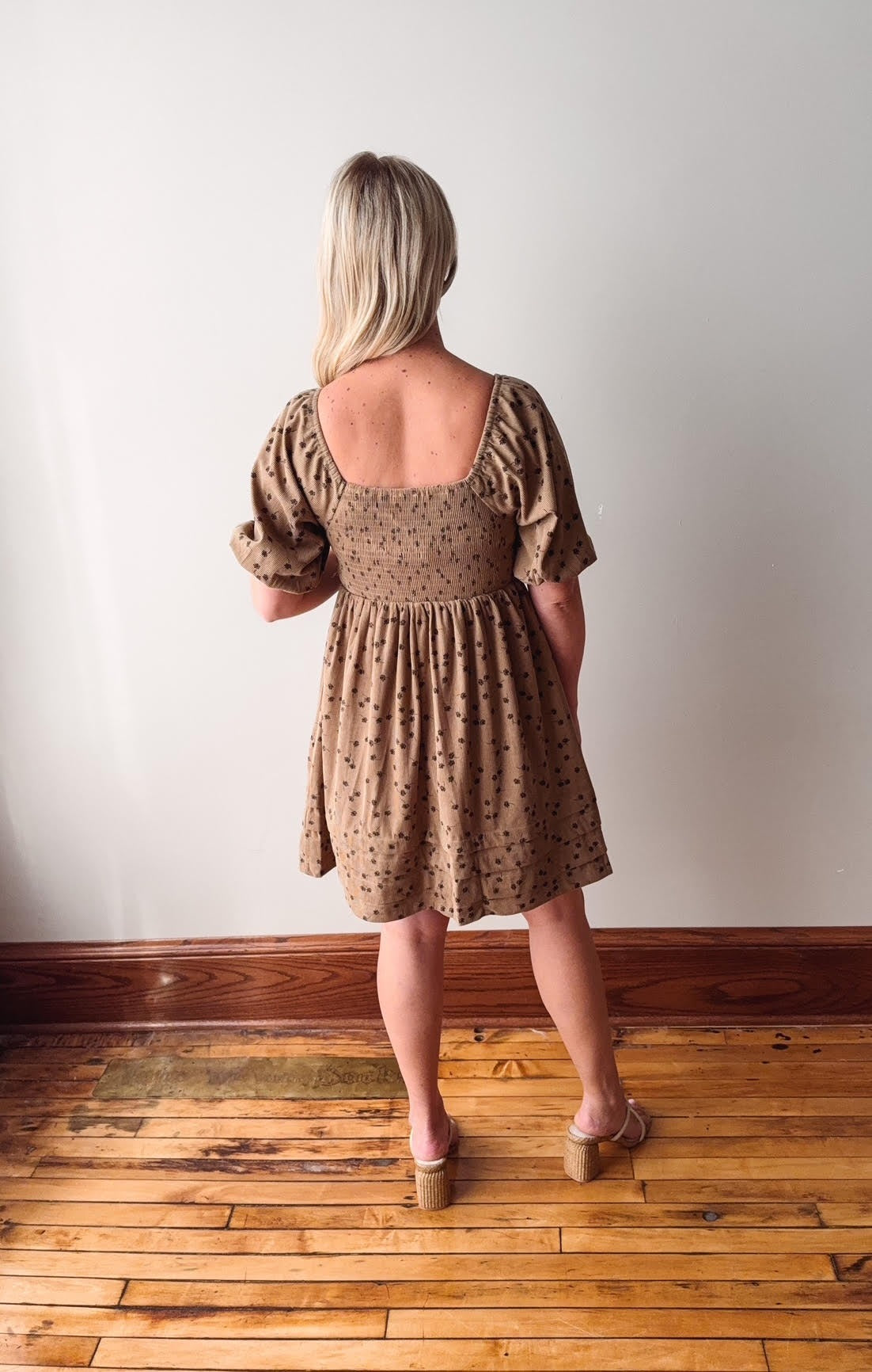 Woman wearing a brown patterned dress standing on a wooden floor with a white wall background