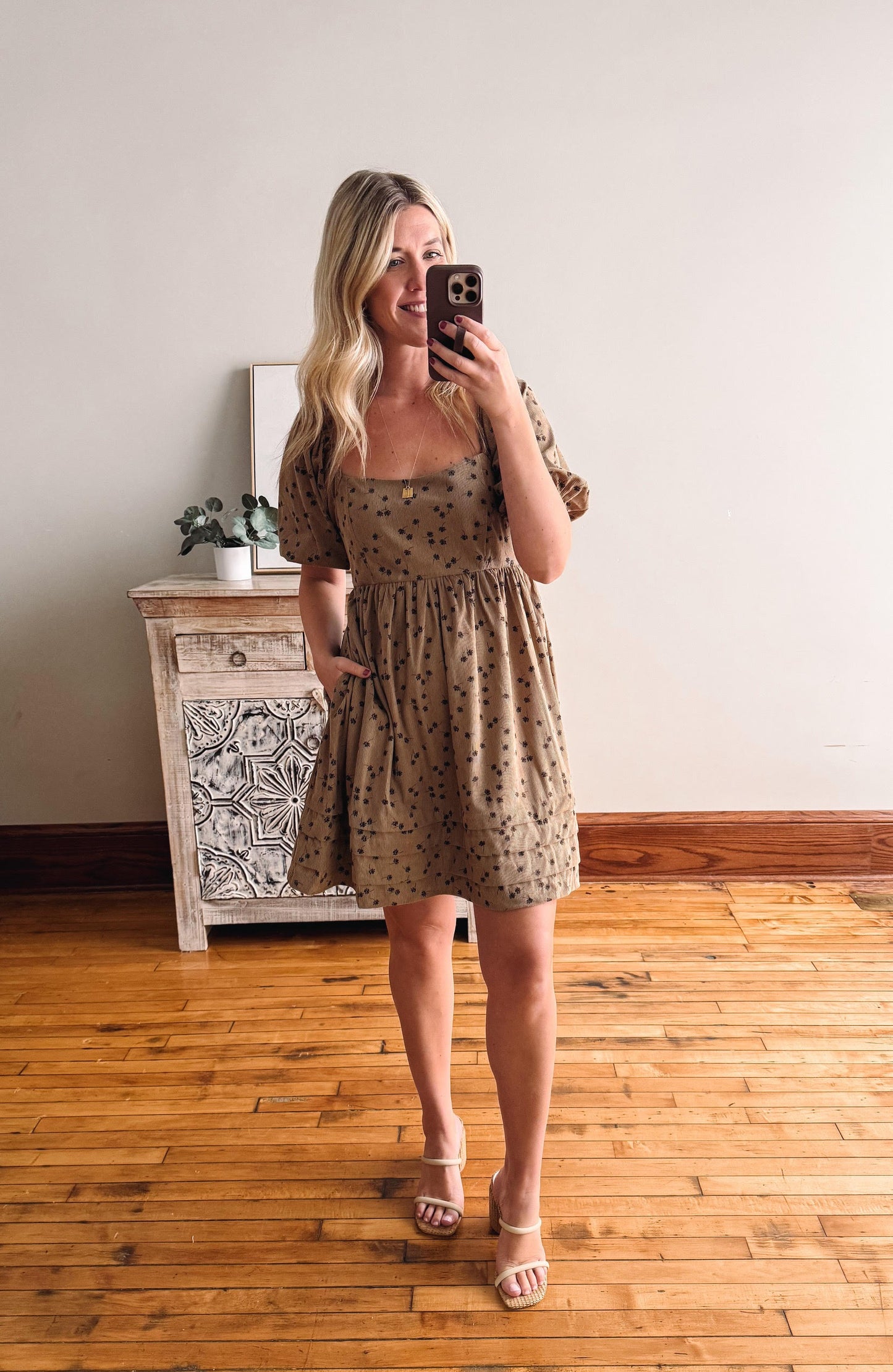 Woman taking a mirror selfie wearing a floral dress in a room with wooden flooring and a decorative cabinet.