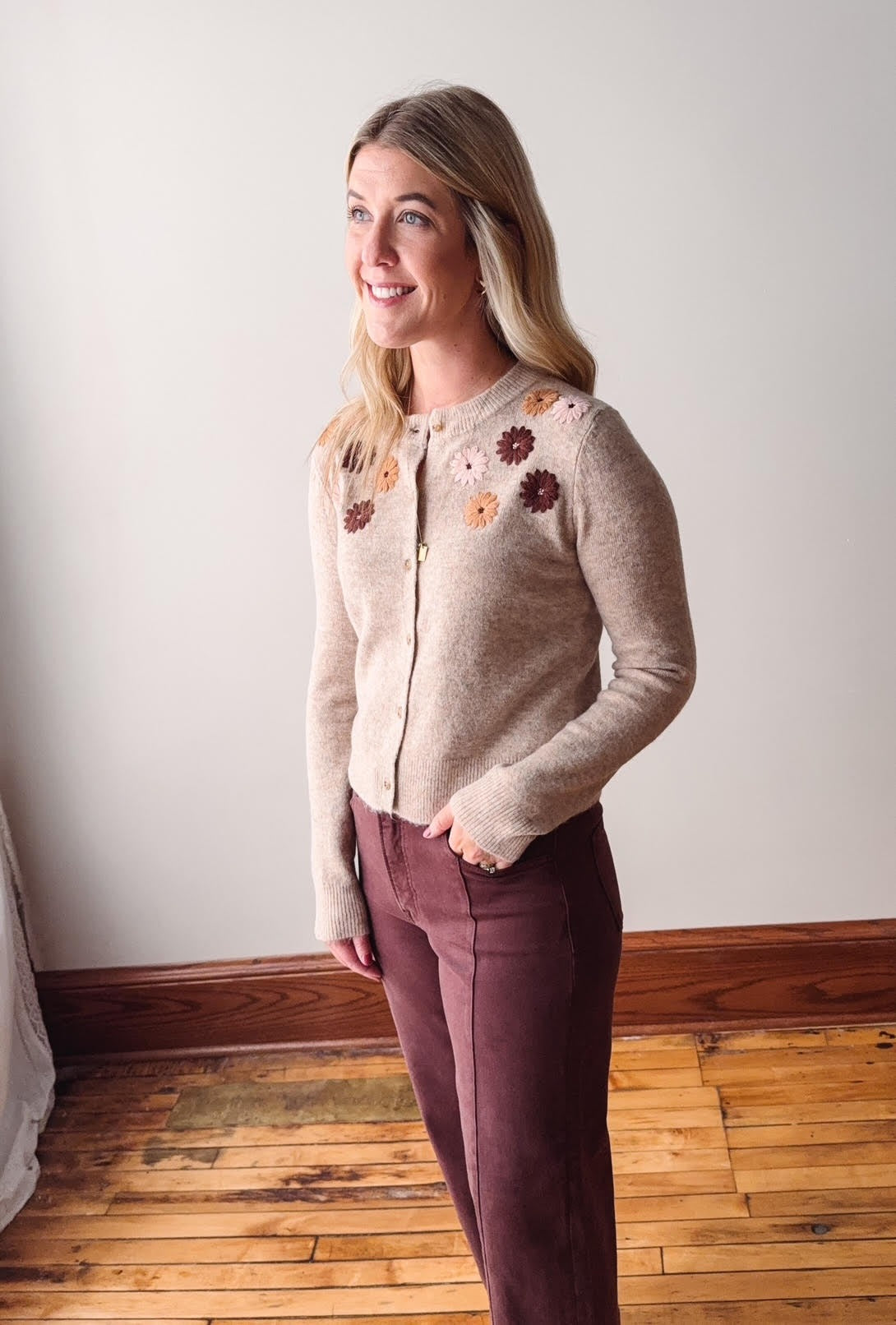 Woman wearing a taupe cardigan with floral embellishments against a white wall.