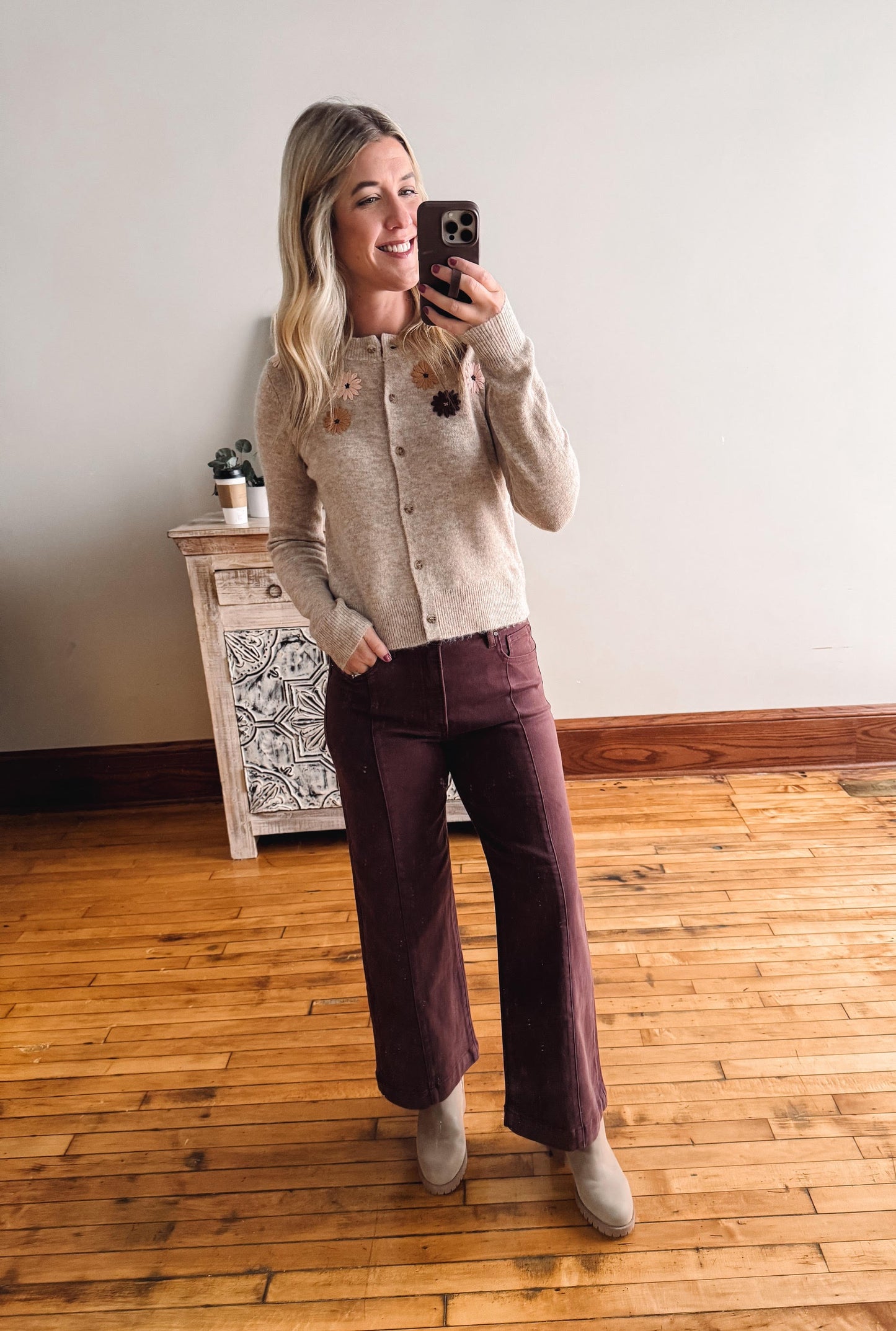 Woman taking a mirror selfie wearing a taupe sweater and maroon pants in a room with wooden flooring.