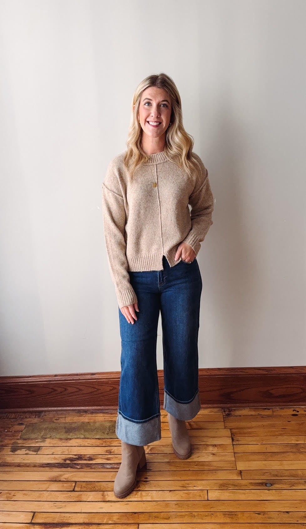 Woman wearing a beige sweater and blue jeans standing on a wooden floor against a white wall.