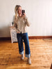 Woman taking a mirror selfie wearing a beige sweater and blue jeans in a room with wooden flooring.