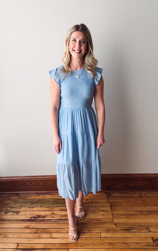 Woman wearing a light blue dress standing on a wooden floor with a white wall background