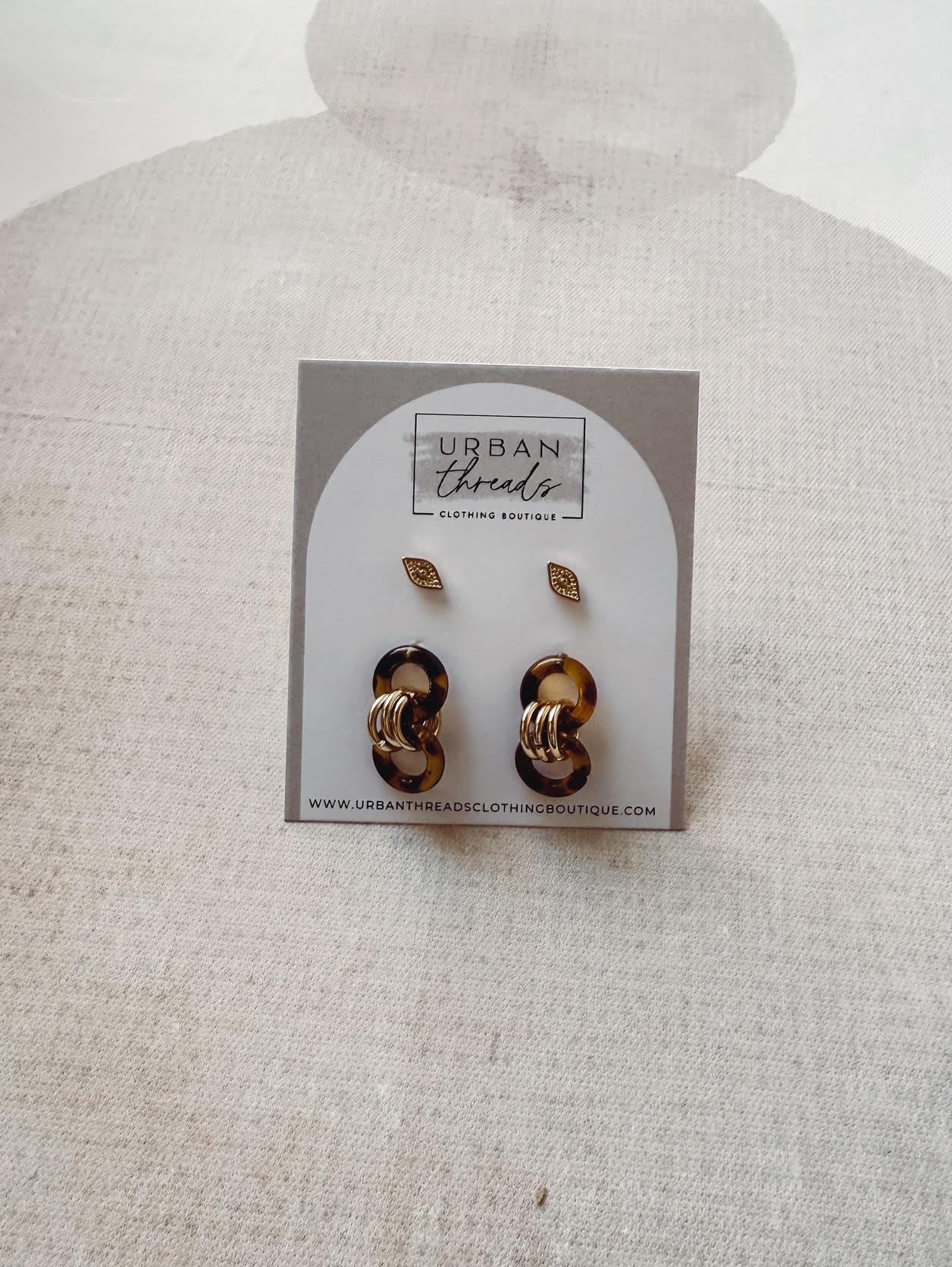 Pair of gold earrings on a card with 'Urban Threads' branding against a beige background