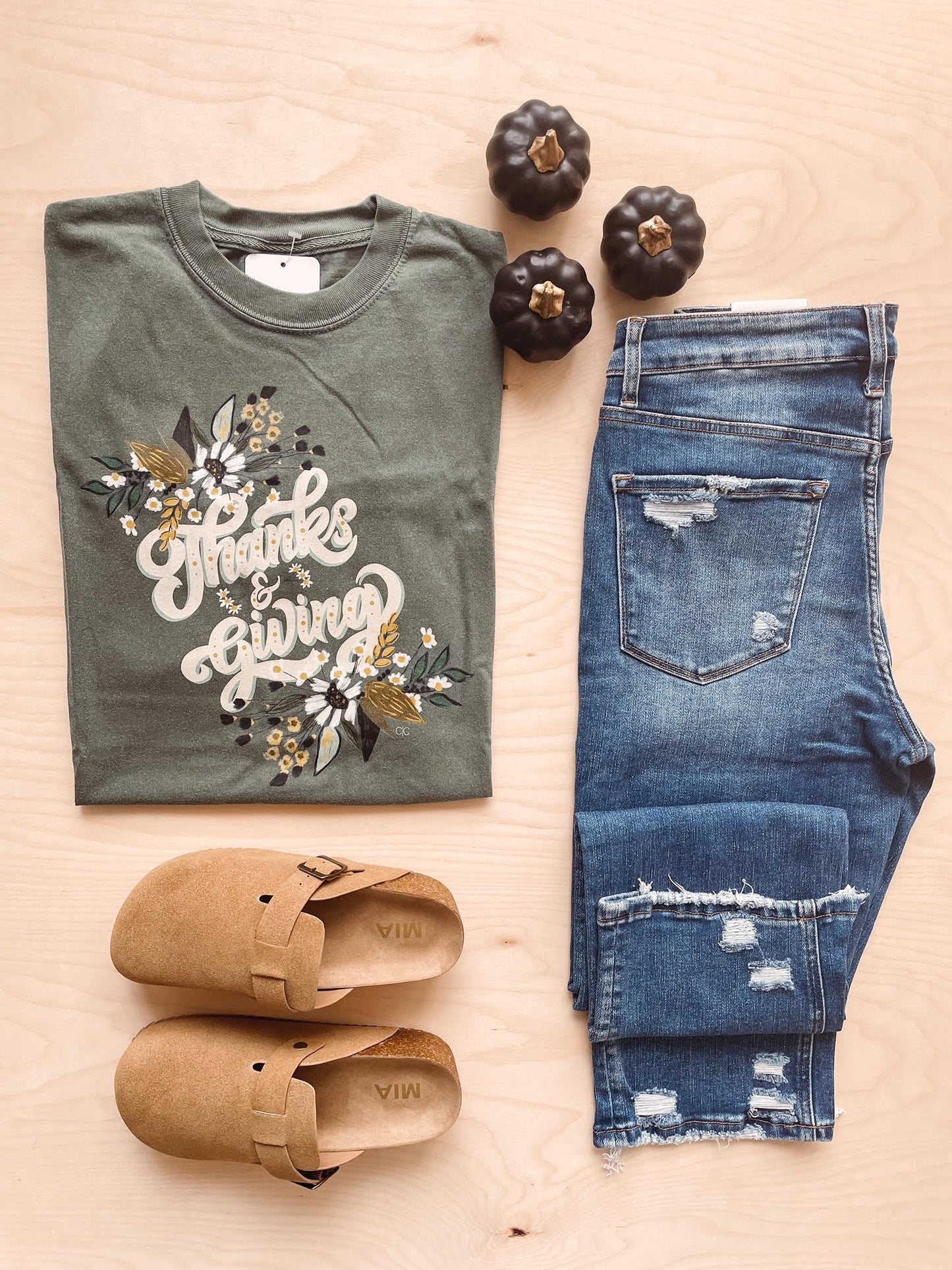 Olive green t-shirt with a floral pattern and text 'Thanks + Giving' laid out flat with blue jeans and brown shoes