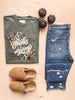 Olive green t-shirt with a floral pattern and text 'Thanks + Giving' laid out flat with blue jeans and brown shoes