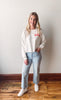 Woman wearing a white sweatshirt with 'USA' and light blue jeans standing against a plain background.