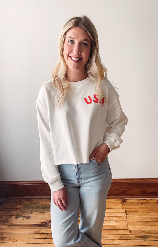 A woman smiling and wearing an off white sweatshirt with 'USA' written in bold red font on the front.
